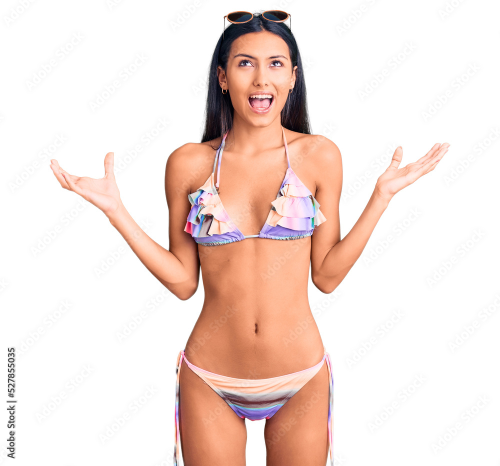Young beautiful latin girl wearing bikini and sunglasses crazy and mad shouting and yelling with aggressive expression and arms raised. frustration concept. Young beautiful latin girl wearing bikini and sunglasses crazy and mad shouting and yelling with aggressive expression and arms raised. frustration concept.