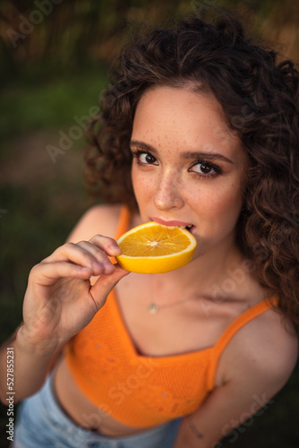 beautiful girl with afro curls eats an orange. .Beautiful portrait of a girl with freckles and curly hair. The girl eats an orange.Corn field. The girl smiles and covers her face with an orange slice