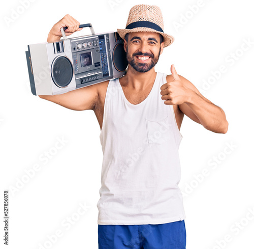 Young hispanic man holding boombox, listening to music smiling happy and positive, thumb up doing excellent and approval sign