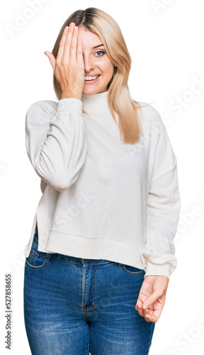 Young caucasian woman wearing casual winter sweater covering one eye with hand, confident smile on face and surprise emotion.