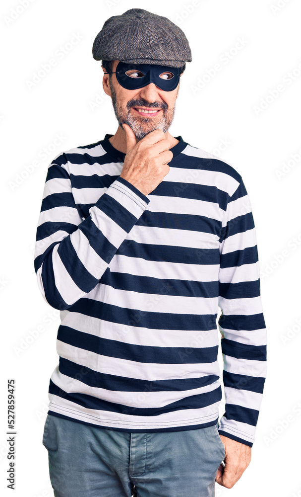 Middle age handsome man wearing burglar mask with hand on chin thinking about question, pensive expression. smiling with thoughtful face. doubt concept. Middle age handsome man wearing burglar mask with hand on chin thinking about question, pensive expression. smiling with thoughtful face. doubt concept.