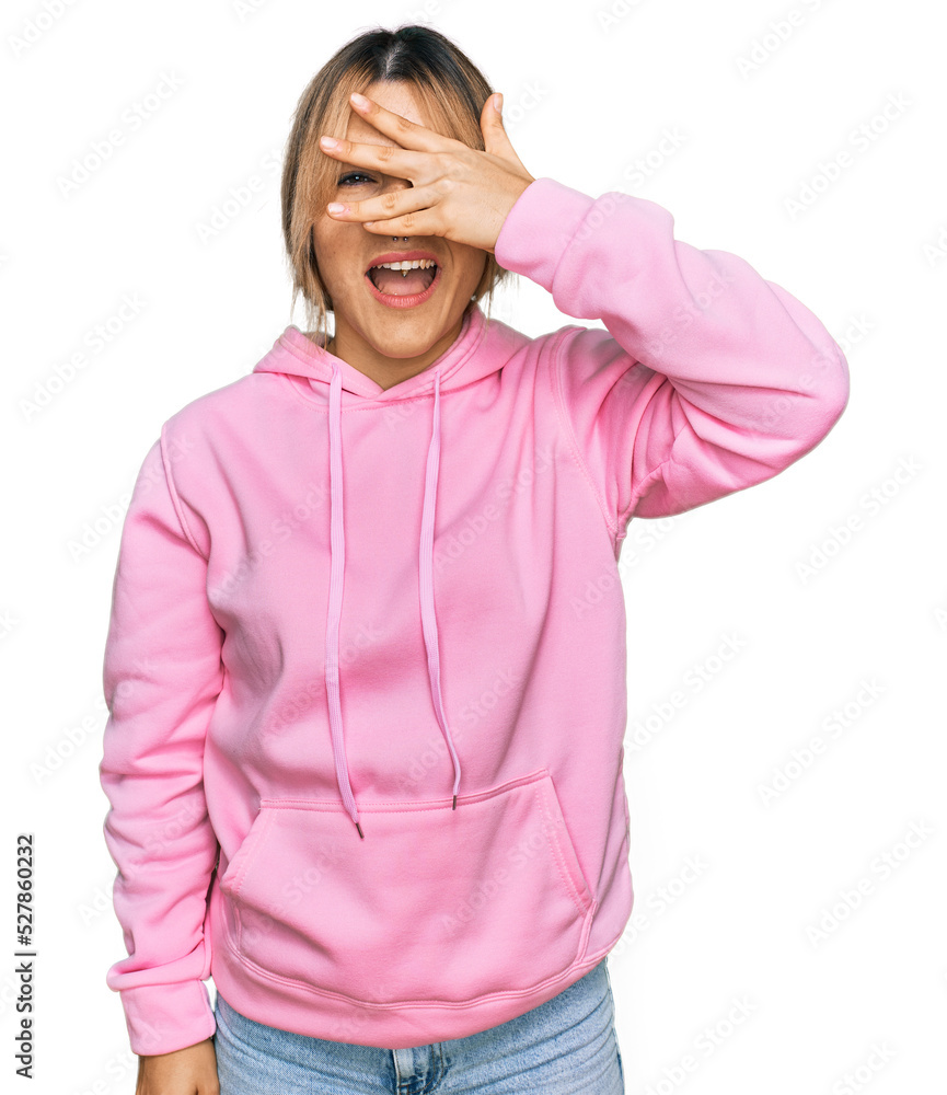 Young caucasian woman wearing casual sweatshirt peeking in shock covering face and eyes with hand, looking through fingers with embarrassed expression. Young caucasian woman wearing casual sweatshirt peeking in shock covering face and eyes with hand, looking through fingers with embarrassed expression.