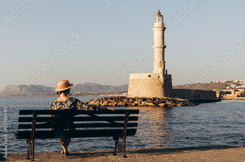 a young woman in a hat sits on the promenade of chania in greece with a lighthouse in the background photo