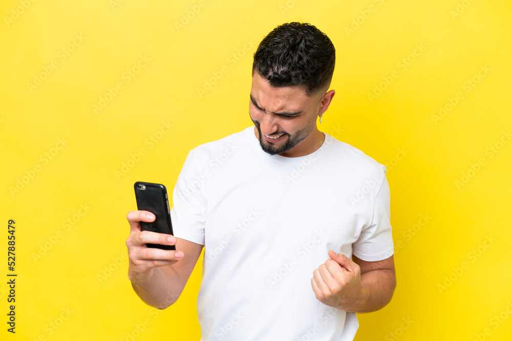 Young Arab handsome man isolated on yellow background using mobile phone and doing victory gesture