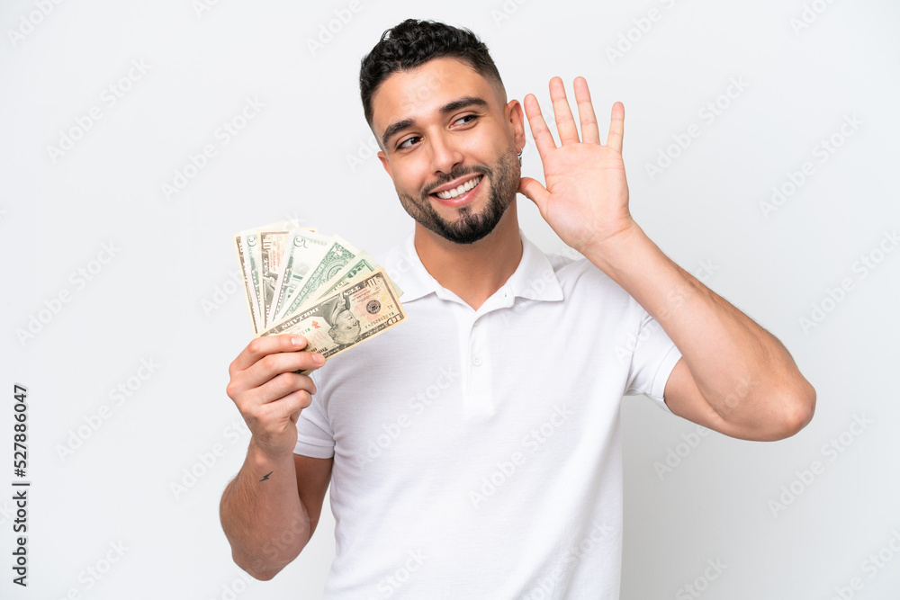 Young Arab man taking a lot of money isolated on white background listening to something by putting hand on the ear Young Arab man taking a lot of money isolated on white background listening to something by putting hand on the ear