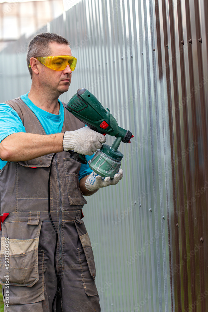 Man in overalls and protective glasses with spray gun in hand. Painting of metal fence Man in overalls and protective glasses with spray gun in hand. Painting of metal fence
