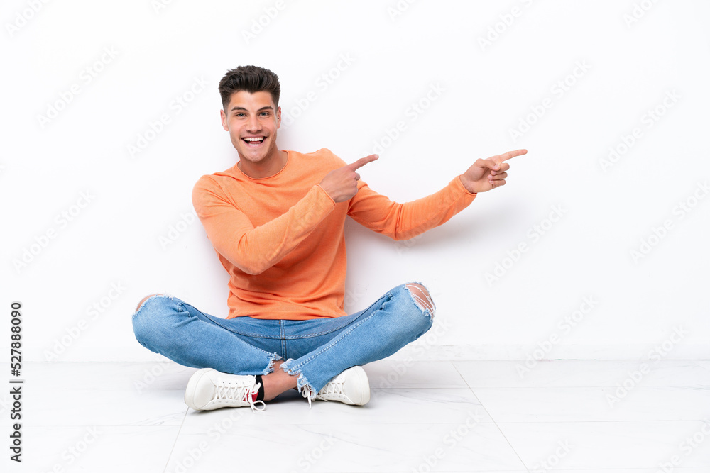 Young man sitting on the floor isolated on white background pointing finger to the side and presenting a product Young man sitting on the floor isolated on white background pointing finger to the side and presenting a product