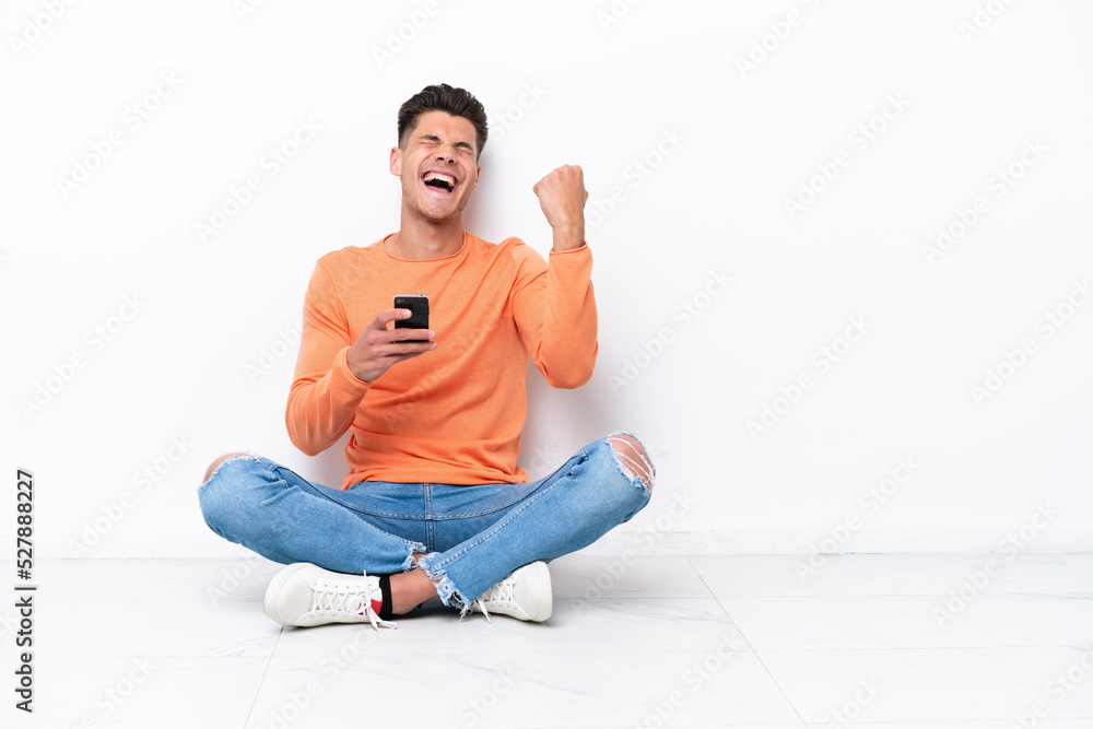 Young man sitting on the floor isolated on white background with phone in victory position Young man sitting on the floor isolated on white background with phone in victory position