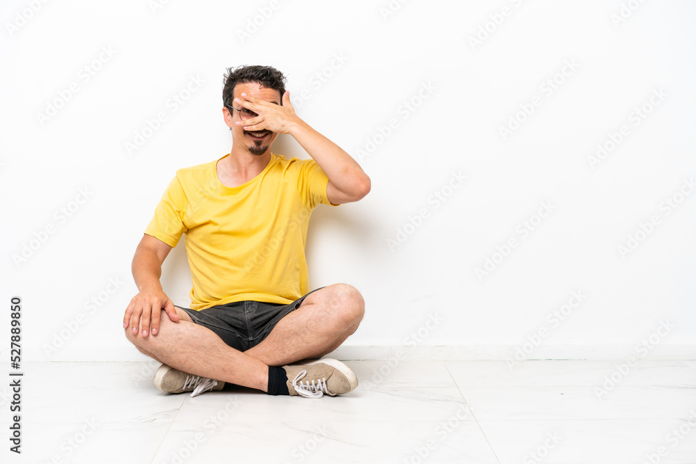Young caucasian man sitting on the floor isolated on white background covering eyes by hands and smiling