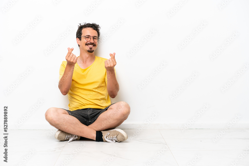 Young caucasian man sitting on the floor isolated on white background making money gesture Young caucasian man sitting on the floor isolated on white background making money gesture