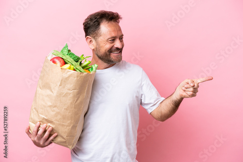 Middle age man holding a grocery shopping bag isolated on pink background pointing finger to the side and presenting a product