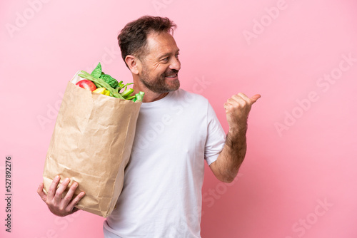 Middle age man holding a grocery shopping bag isolated on pink background pointing to the side to present a product