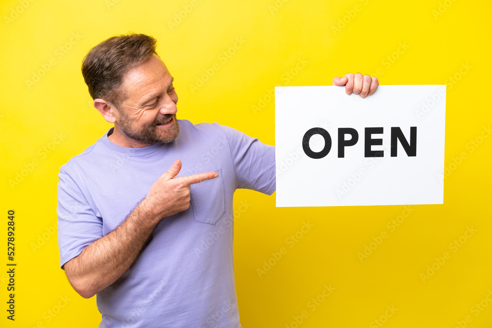 Middle age caucasian man isolated on yellow background holding a placard with text OPEN and pointing it Middle age caucasian man isolated on yellow background holding a placard with text OPEN and pointing it