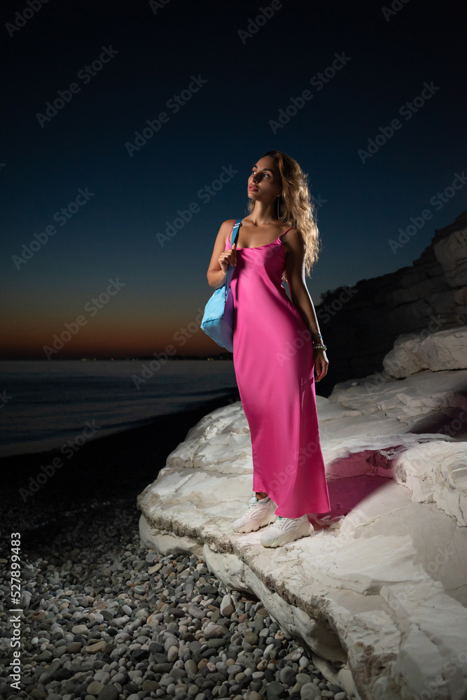 a beautiful stylish girl with a backpack in a fashionable pink dress on the seashore among the rocks at sunset