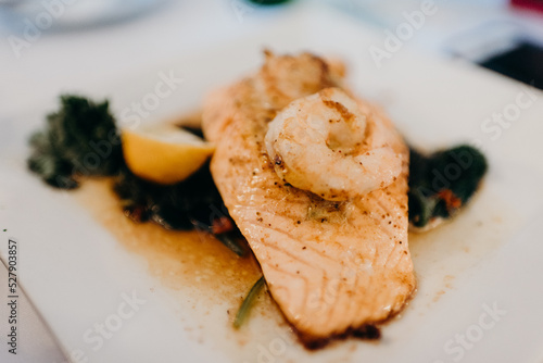 Piece of salmon with piece of shrimp resting on top over a bed of greens and lemon wedge
