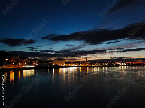 Night promenade in the distance by the river with illuminated houses. On the background of a dark blue sky with clouds © Serhii