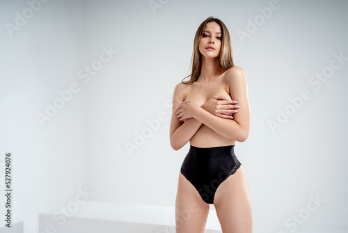 Photo of a beautiful sexy female model with slim body. Sensual woman posing in black panties, covering breast with hands, looking at the camera.