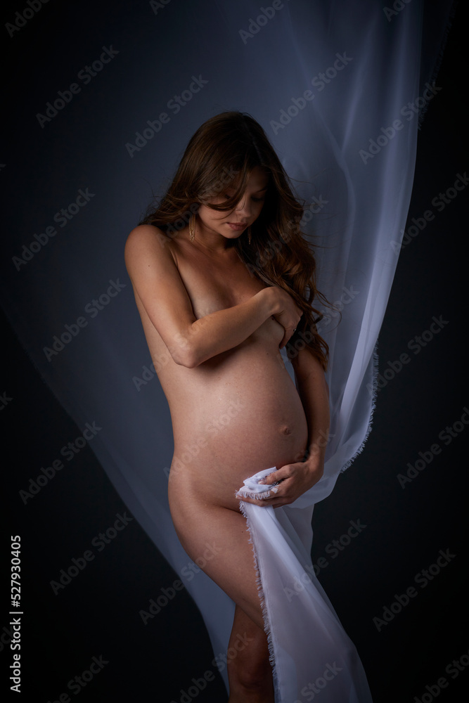 Naklejka premium Beautiful young pregnant serious sexy woman with long curly hair and big belly, without clothes sitting in dark studio and covers her breasts with her hand