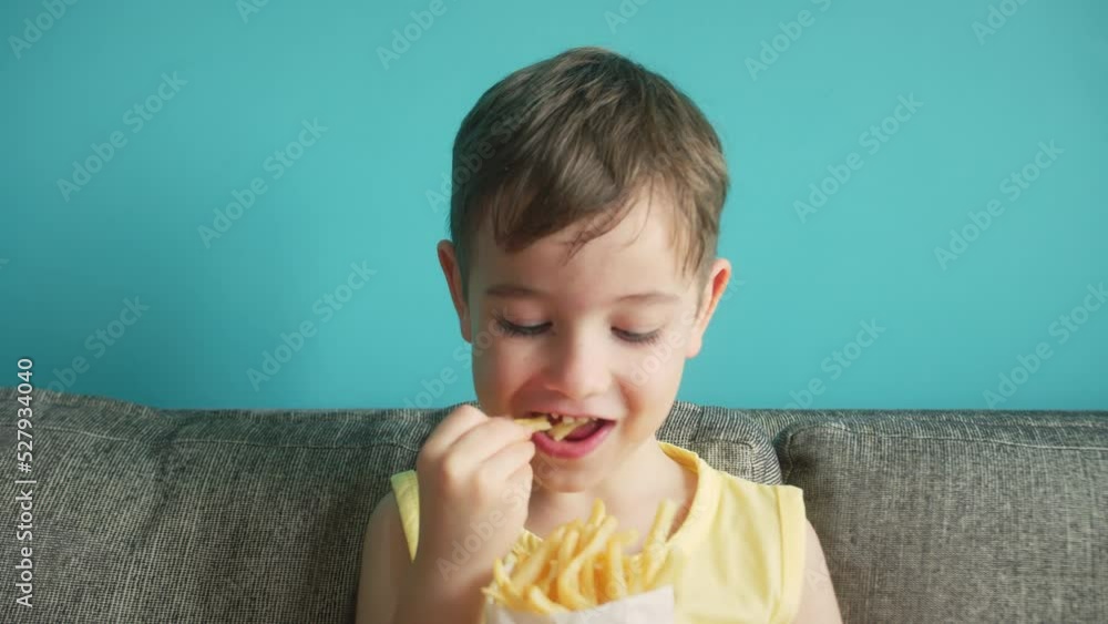 Child eating french fries on blue background. Cute little boy licks her fingers eating a potato french fries. Hunny Baby eat fast food. little girl happily eating fried lifestyle potatoes Child eating french fries on blue background. Cute little boy licks her fingers eating a potato french fries. Hunny Baby eat fast food. little girl happily eating fried lifestyle potatoes