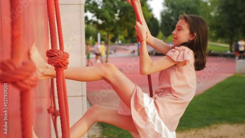 A little girl climbs a rope resting her feet on special ledges on a children's climbing wall in the park.