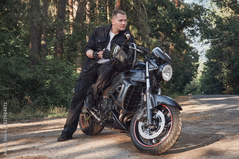 Fototapeta premium Young male biker travels on a motorcycle alone, stopped and using a mobile phone on the side of a forest road