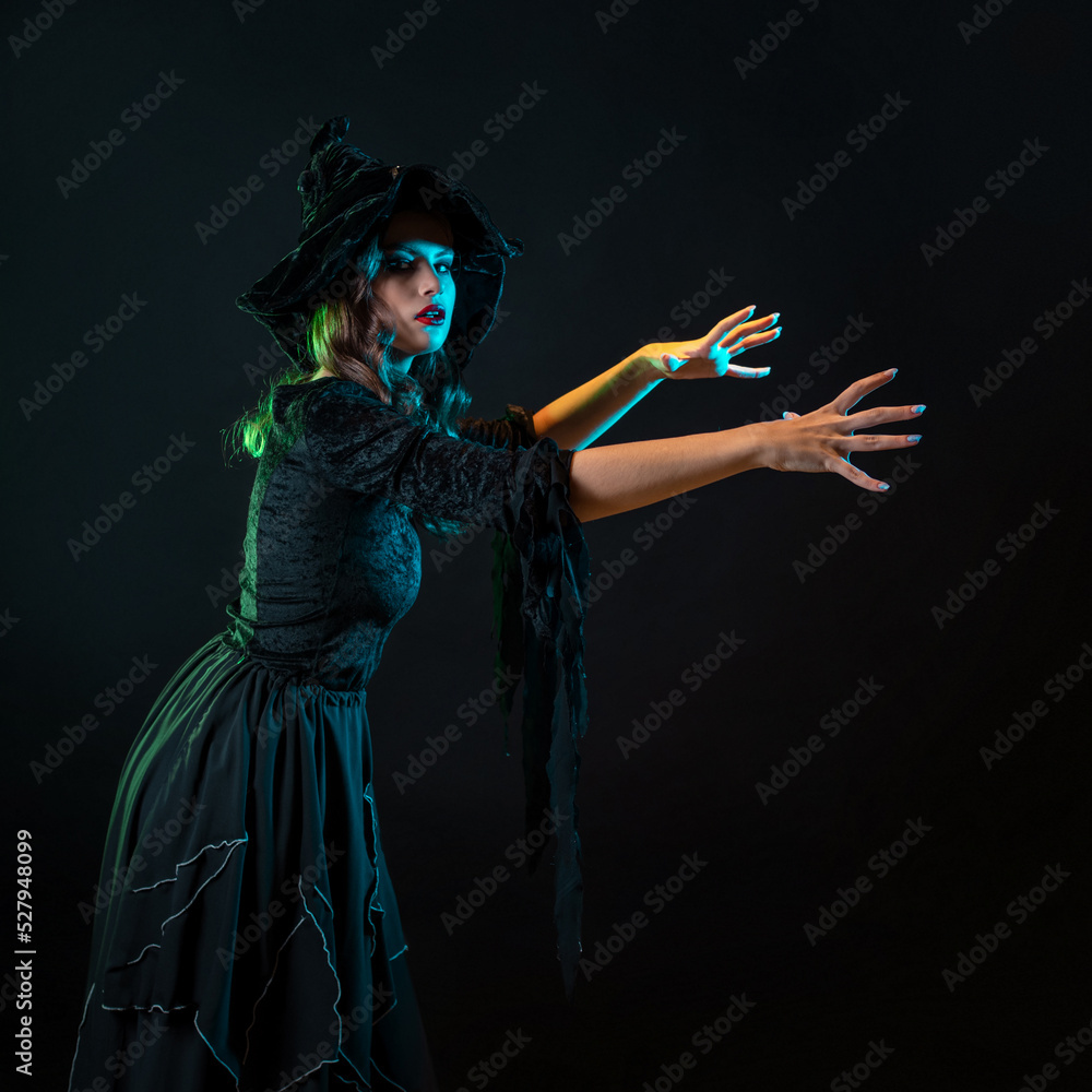 A charming young witch in a costume makes magical passes with two hands, a caster. Young brunette in pointed hat and black dress, photo on black