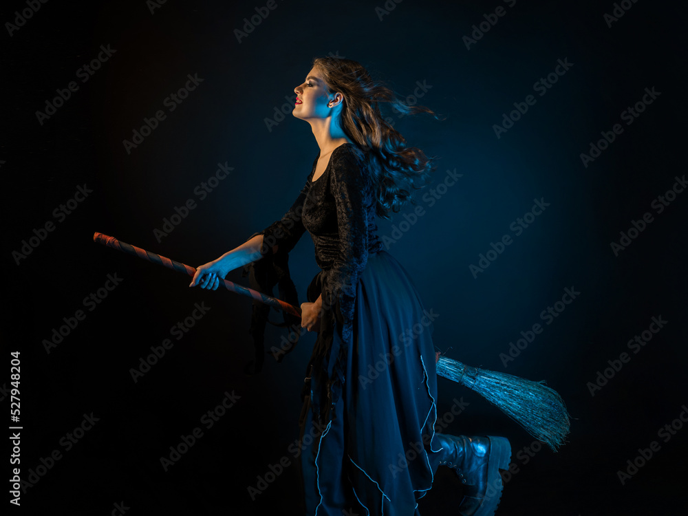 A beautiful witch on a broomstick is flying to the sabbath. Young brunette woman in a dress and with a broom, rushing to a Halloween party, fire lighting, black background