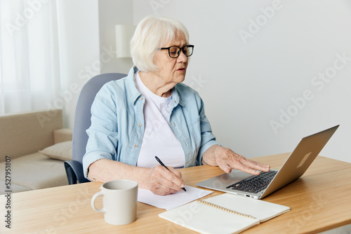 an attentive, serious elderly woman with white gray hair is sitting in a light blue shirt and a white T-shirt at home at her desk with a laptop and a notepad for notes