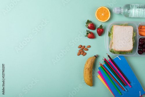 Fruits with sandwich in tiffin box by water bottle, color pencils and books on green background