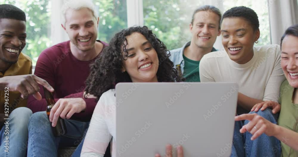 Diverse group of happy male and female friends looking at laptop and laughing in living room Diverse group of happy male and female friends looking at laptop and laughing in living room