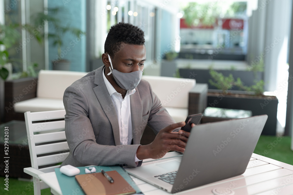 African american male businessman sitting in a cafe wearing face mask using smartphone and laptop African american male businessman sitting in a cafe wearing face mask using smartphone and laptop