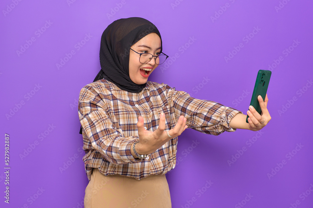 Shocked young Asian woman in plaid shirt using a mobile phone, getting great news isolated on purple background Shocked young Asian woman in plaid shirt using a mobile phone, getting great news isolated on purple background