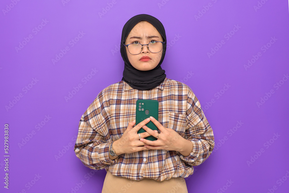 Annoyed young Asian woman in plaid shirt holding a mobile phone, looking at camera isolated on purple background Annoyed young Asian woman in plaid shirt holding a mobile phone, looking at camera isolated on purple background