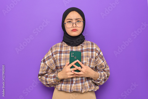 Annoyed young Asian woman in plaid shirt holding a mobile phone, looking at camera isolated on purple background