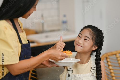 Asian preteen girl showing thumb up when her mom feeding a piece of pizza.