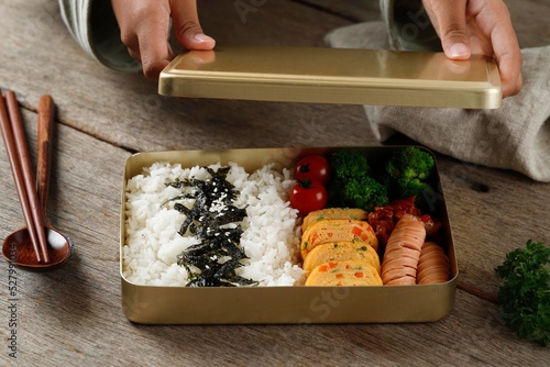Female Hand Preparing Dosirak or Korean Bento Box, Served on Gold  Metal Lunchbox to Keep Food Warm. photo