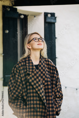 Stylish woman in glasses and coat stnads near house in village