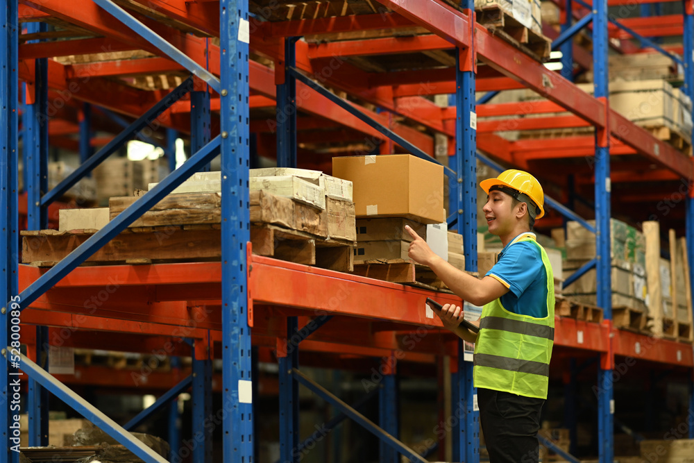 Young man warehouse worker wearing safety hardhat and vest checking quantity of storage product on shelf full of packed boxes