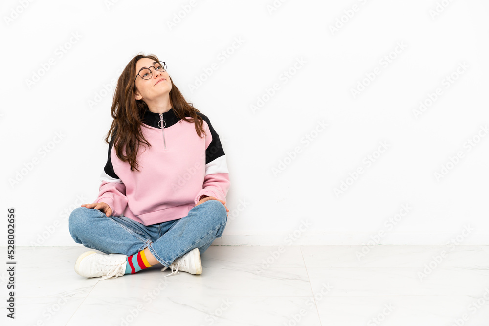 Young caucasian woman sitting on the floor isolated on white background looking up while smiling Young caucasian woman sitting on the floor isolated on white background looking up while smiling