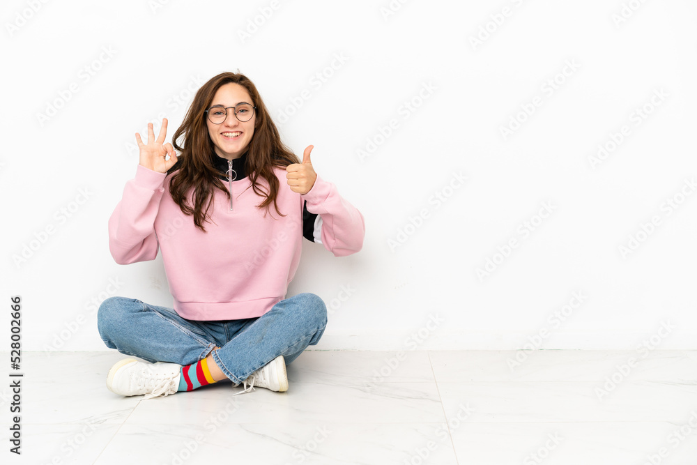 Young caucasian woman sitting on the floor isolated on white background showing ok sign and thumb up gesture Young caucasian woman sitting on the floor isolated on white background showing ok sign and thumb up gesture