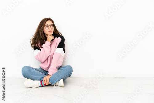 Young caucasian woman sitting on the floor isolated on white background having doubts and with confuse face expression