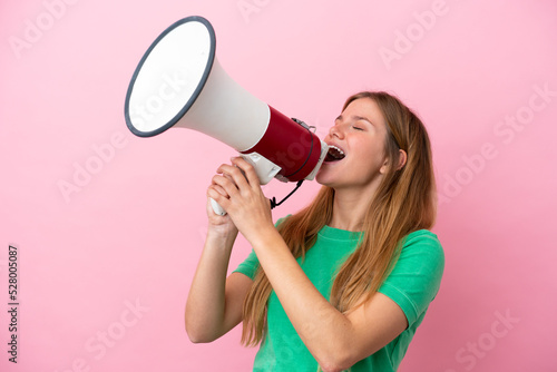 Young blonde woman isolated on pink background shouting through a megaphone to announce something in lateral position