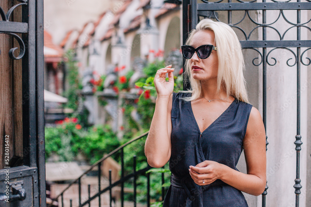 Fototapeta premium A young, blonde girl in a black blouse and sunglasses poses against the backdrop of an old courtyard in Lviv. Ukraine.