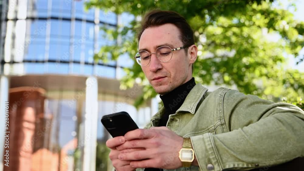 Focusing white man using digital smartphone sit on parapet outside, young Caucasian guy holding telephone looking at screen reading e-book app online relaxing, trading currency online. Focusing white man using digital smartphone sit on parapet outside, young Caucasian guy holding telephone looking at screen reading e-book app online relaxing, trading currency online.