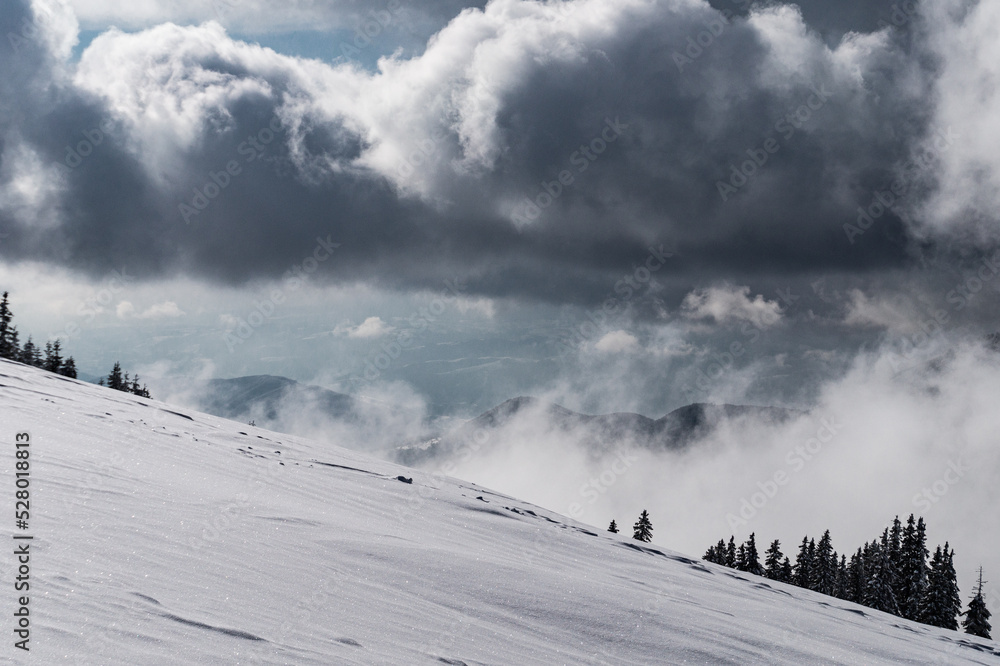 Winter landscape. Dramatic sky with grey clouds above snow covered steep mountain slope. Untouched white powder snow contrasts the grey clouds.