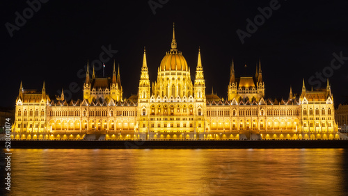 Panorama of the Hungarian Parliament building at night. Night view of the illuminated main facade. Warm lights reflected in the water. Situated in Budapest on the eastern bank of the Danube.