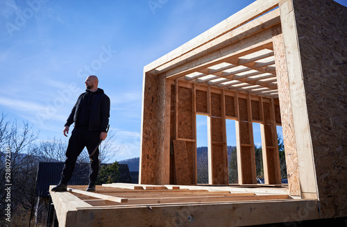 Wallpaper Mural Male builder building wooden frame house. Bald man standing on construction site, inspecting quality of work on sunny day with blue sky on background. Torontodigital.ca