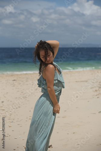 A beautiful woman in a blue dress and with dark curly hair is having fun on the beach. Tourist vacation in a tropical resort. A cheerful girl with a curly hairstyle. Free lifestyle.