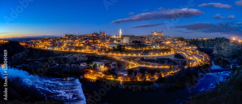 Panoramic view of the city of Toledo with the streetlights on, illuminating the streets and famous buildings, such as the Cathedral, along the Tagus River at dusk
