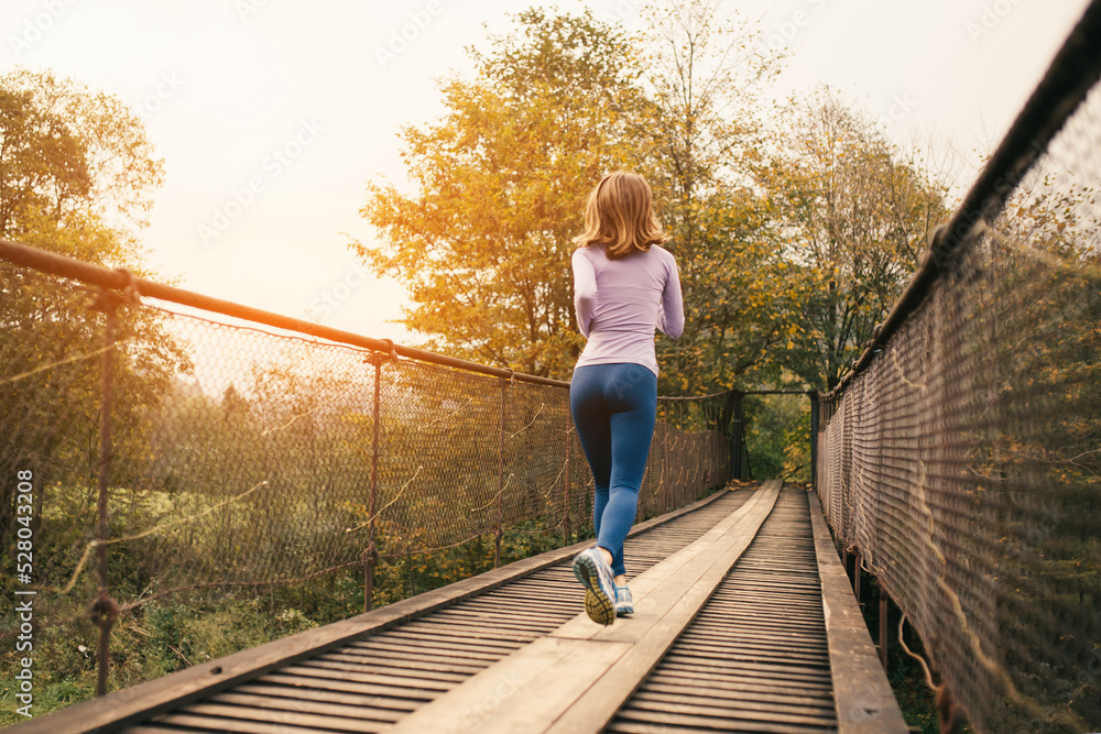 Rear view of a sporty woman runner running over an old suspension bridge on a cool autumn day.